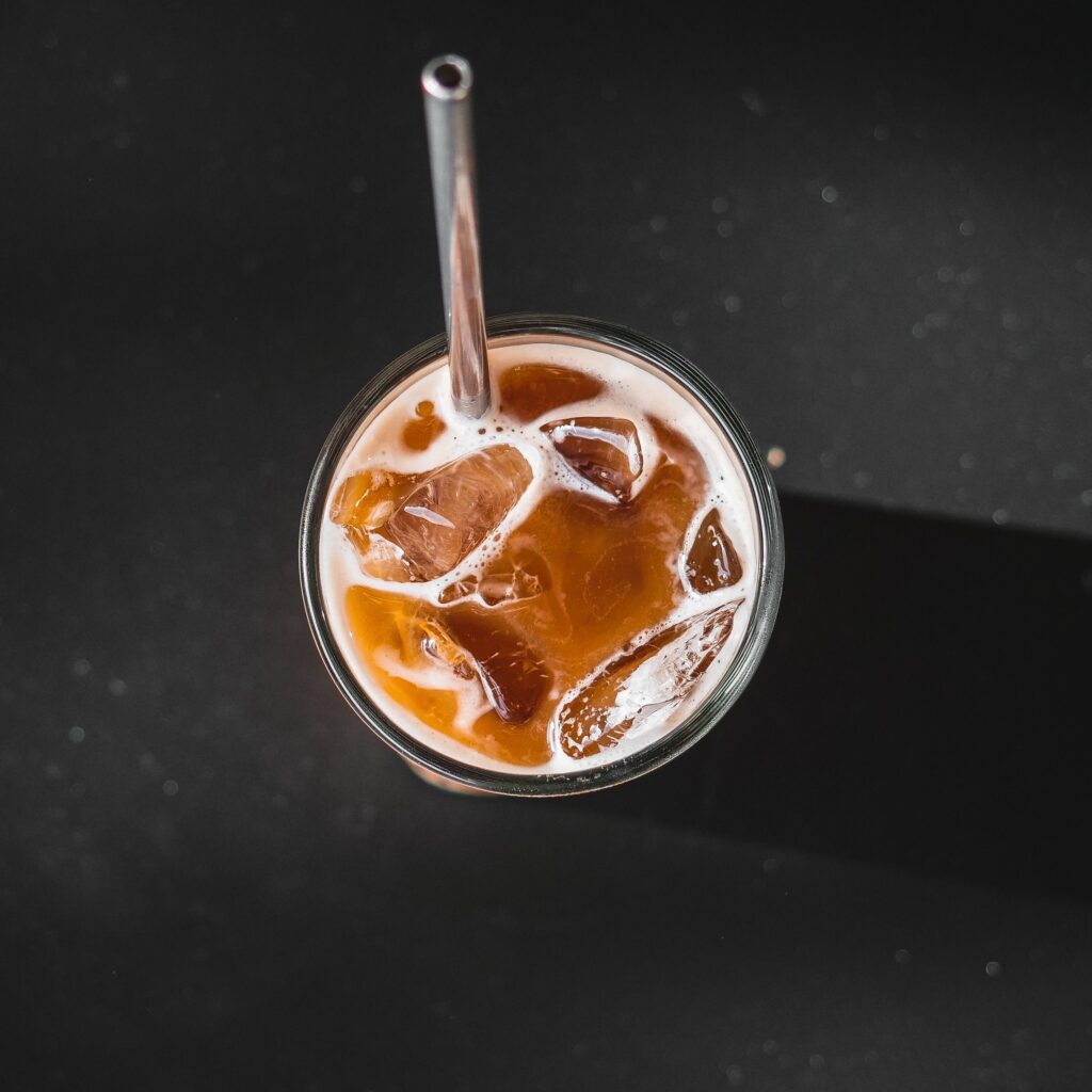 Top view of a glass filled with iced coffee and a metal straw, casting shadows on a dark background.